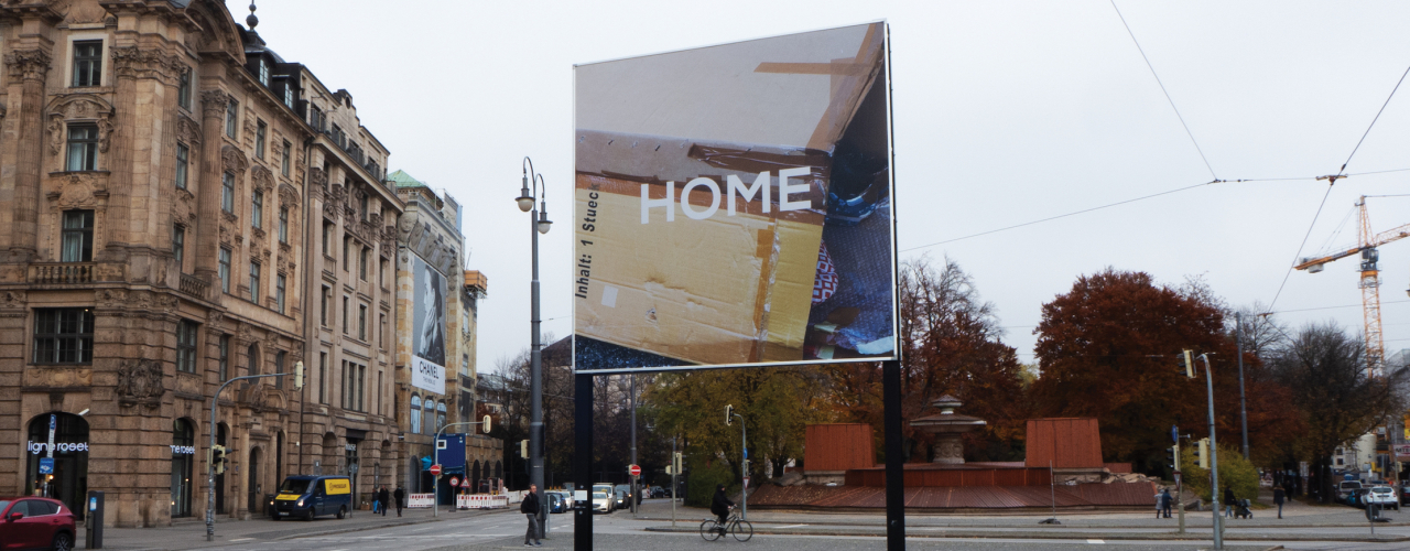 Diagonal view of the billboard. The motif shows cardboard boxes glued together to form a temporary canopy for housing.