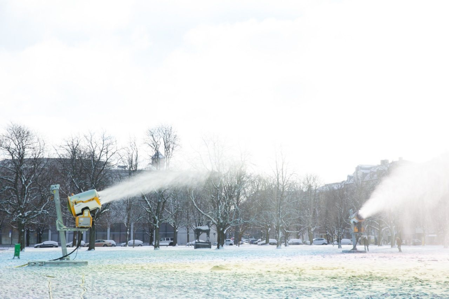 View of two snow cannons spraying coloured snow onto the lawn in front of Alte Pinakothek. In the background is a row of trees.