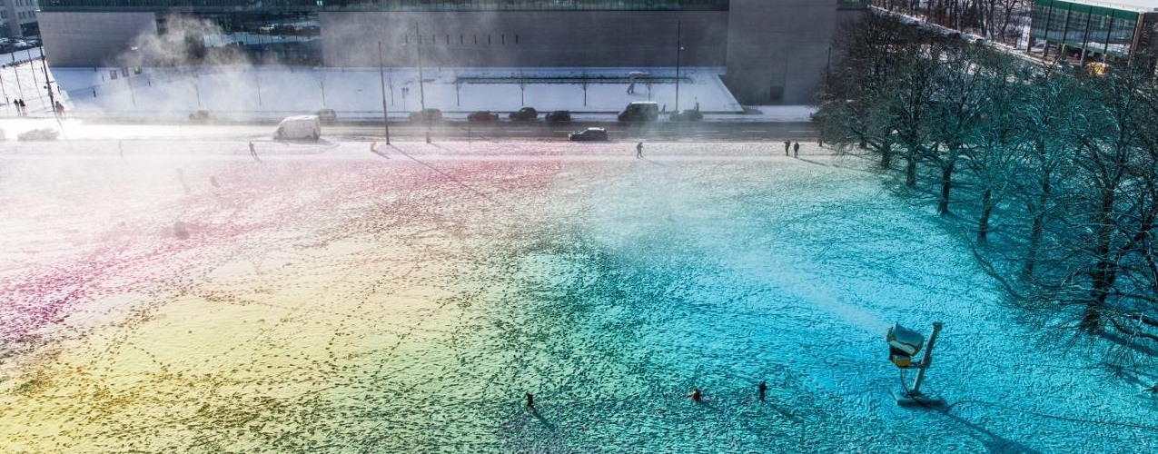 Blick von oben auf eine surreale bunte Schneefläche mit einer laufenden Schneekanone. Im Hintergrund zu sehen ist die Hochschule für Film und Fernsehen.