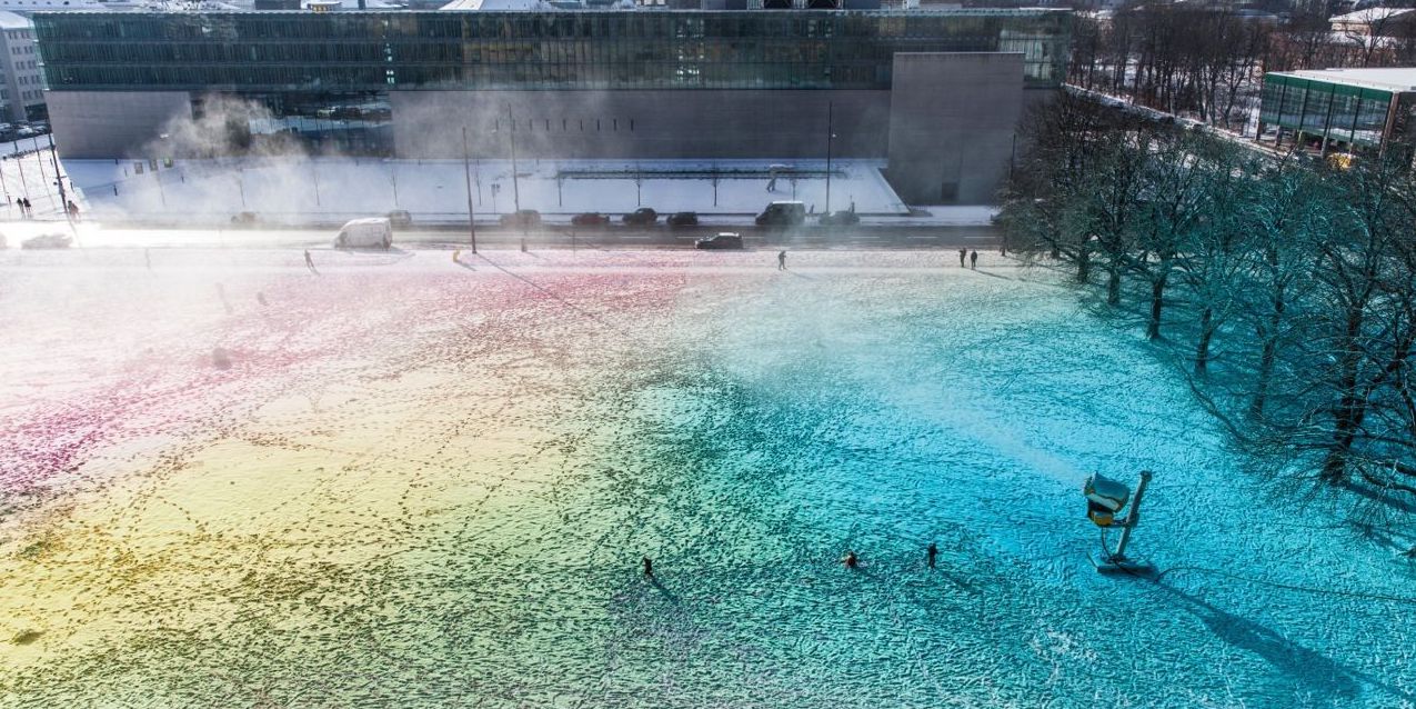Blick von oben auf eine surreale bunte Schneefläche mit einer laufenden Schneekanone. Im Hintergrund zu sehen ist die Hochschule für Film und Fernsehen.