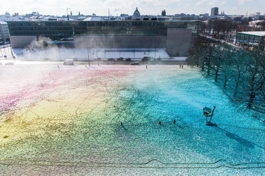 Blick von oben auf eine surreale bunte Schneefläche mit einer laufenden Schneekanone. Im Hintergrund zu sehen ist die Hochschule für Film und Fernsehen.