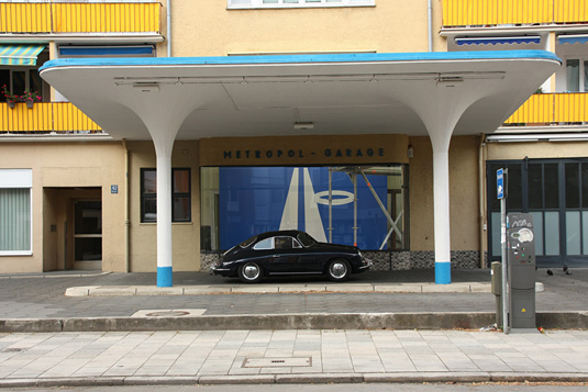 Ein schwarzer Porsche-Oldtimer ist unter dem weiß-blauen Vordach der ehemaligen Metropol Tankstelle geparkt.