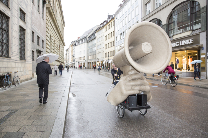 The picture shows a sculpture in the form of a giant megaphone held by two oversized hands. The sculpture is mounted on a bicycle together with a sound system and is driven through Residenzstraße in Munich.
