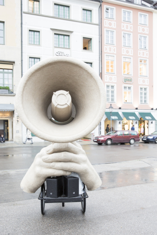 Das Bild zeigt eine Skulptur in Form eines riesigen Megafons, das von zwei überdimensionierten Händen gehalten wird. Die Skulptur ist gemeinsam mit einer Soundanlage auf ein Fahrrad montiert.