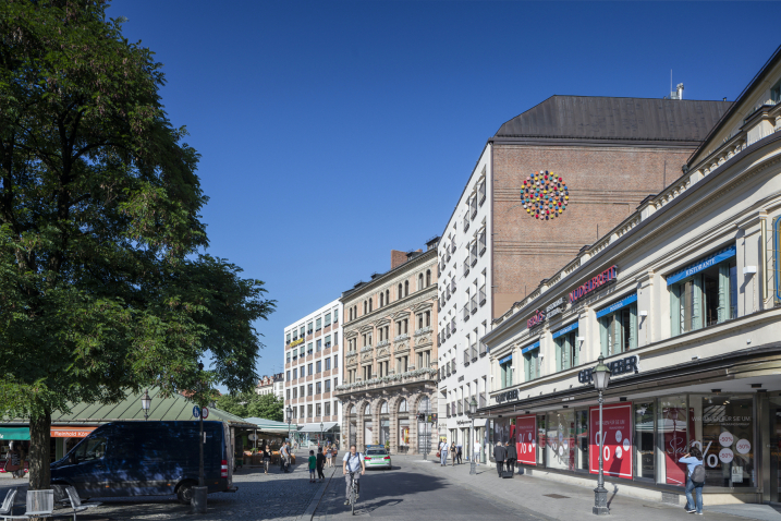 Das Bild zeigt eine Straßenansicht des Viktualienmarkts. Auf der seitlichen Backsteinfassade des Kustermanngebäudes erscheint ein kreisrundes Wandobjekt aus verschiedenfarbig glasierten Keramikscheiben.