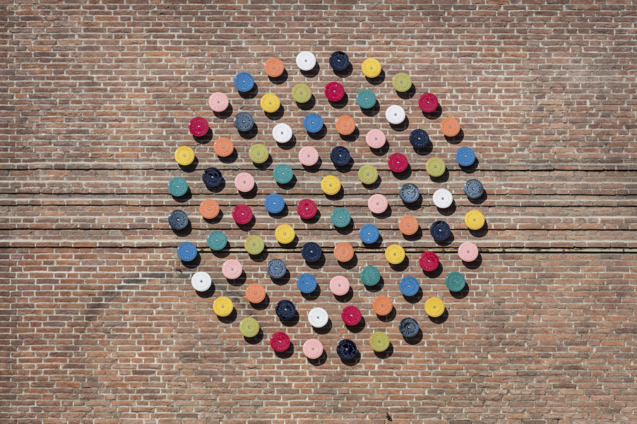 The picture shows 79 differently coloured glazed ceramic discs arranged in a circle on a brick wall.