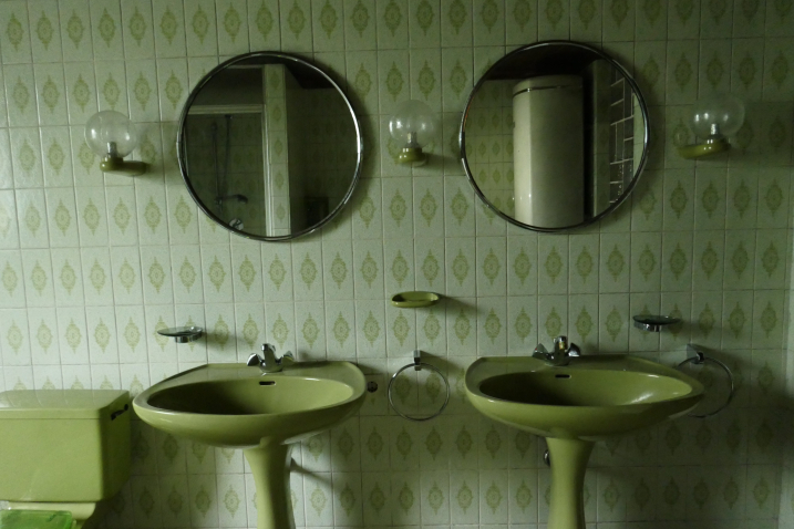 View of a green-tiled bathroom with two green washbasins in the centre and two round mirrors hanging above.