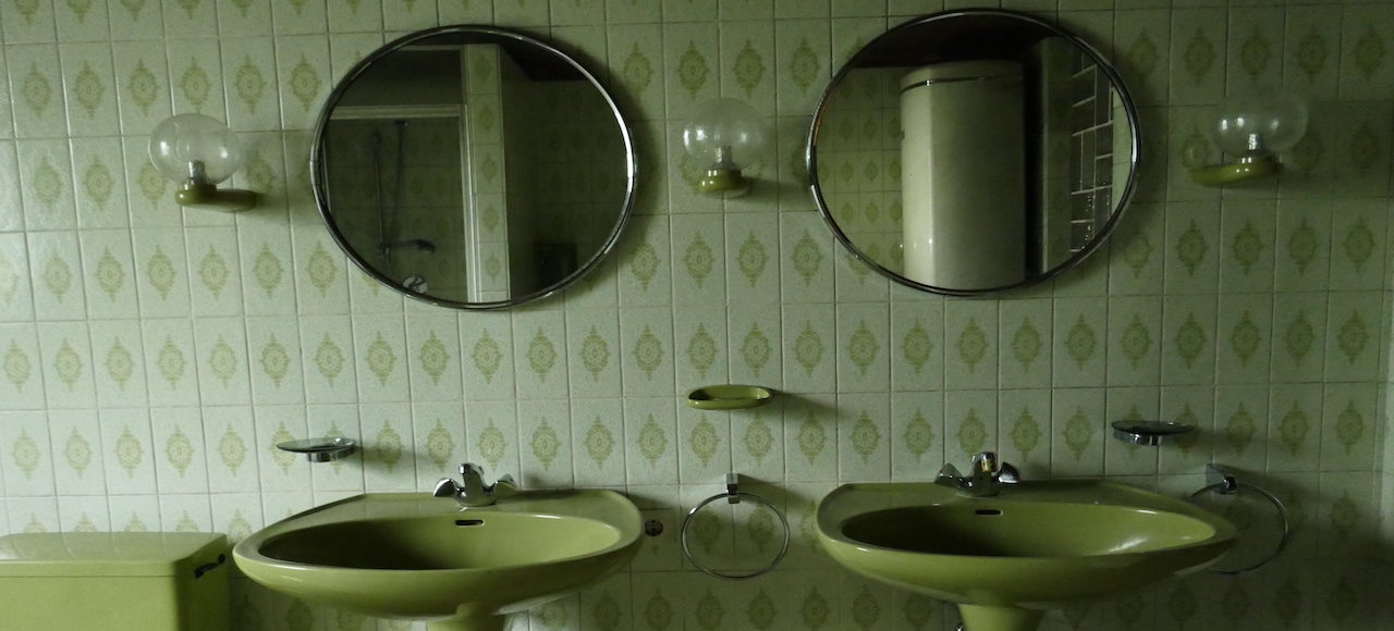 Blick in ein grüngefliestes Badezimmer, in der Mitte zwei grüne Waschbecken sowie zwei darüber hängende runde Spiegel.