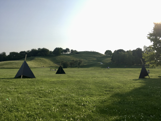 On the lawn in front of the Olympiaberg there are three black wooden triangles for seating, in the background you can see people on the lawn and the Olympiaberg.