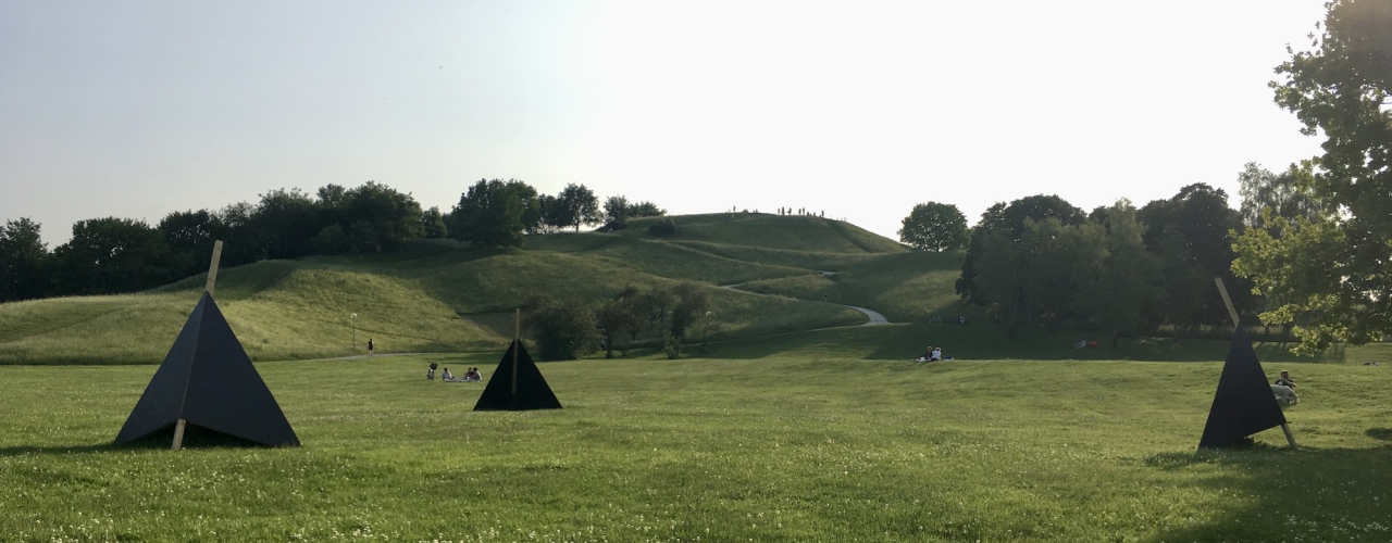 On the lawn in front of the Olympiaberg there are three black wooden triangles for seating, in the background you can see people on the lawn and the Olympiaberg.