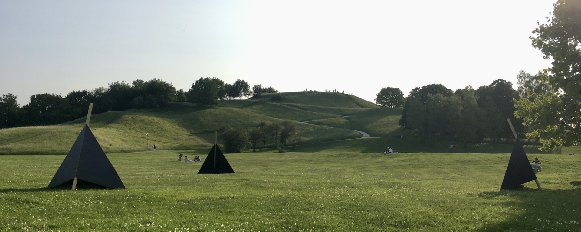 On the lawn in front of the Olympiaberg there are three black wooden triangles for seating, in the background you can see people on the lawn and the Olympiaberg.