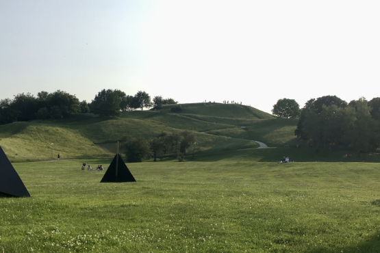 On the lawn in front of the Olympiaberg there are three black wooden triangles for seating, in the background you can see people on the lawn and the Olympiaberg.