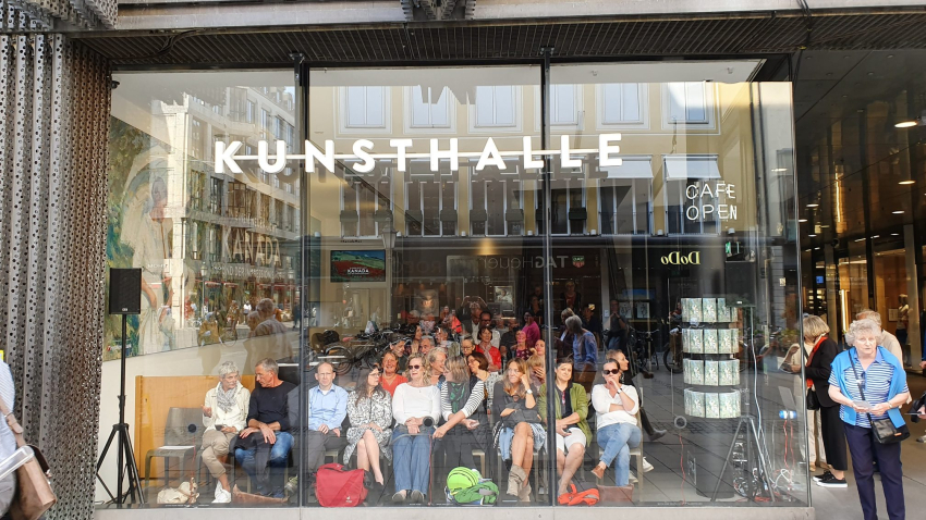 Behind the glass window front of the entrance area of the Kunsthalle München, an audience sits on rows of chairs. The people look through the window onto Theatinerstrasse.