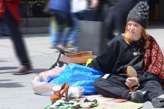 A woman in black clothing and a cap sits on the ground in a pedestrian zone, surrounded by a few belongings. Passers-by walk around her.