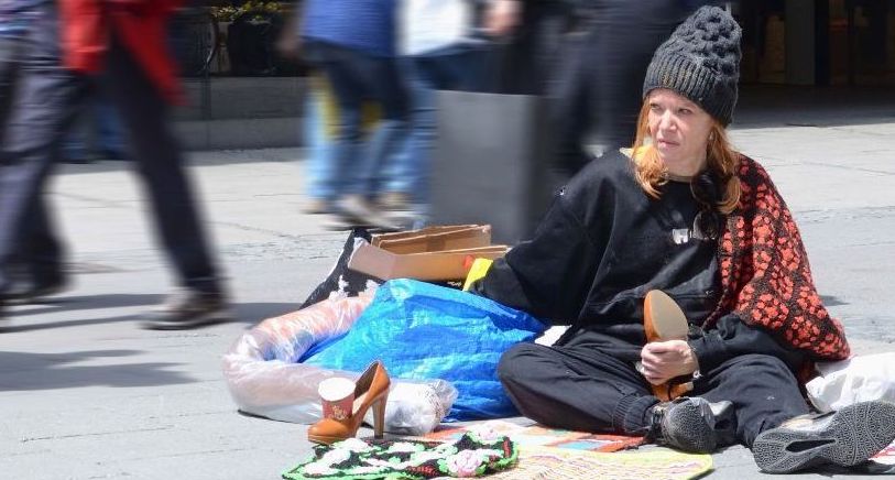 A woman in black clothing and a cap sits on the ground in a pedestrian zone, surrounded by a few belongings. Passers-by walk around her.