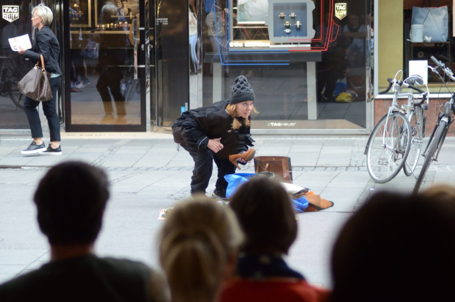 A woman in black clothing and a cap kneels on the ground in a pedestrian zone, surrounded by a few belongings. The heads of a number of people can be seen from behind in the foreground.