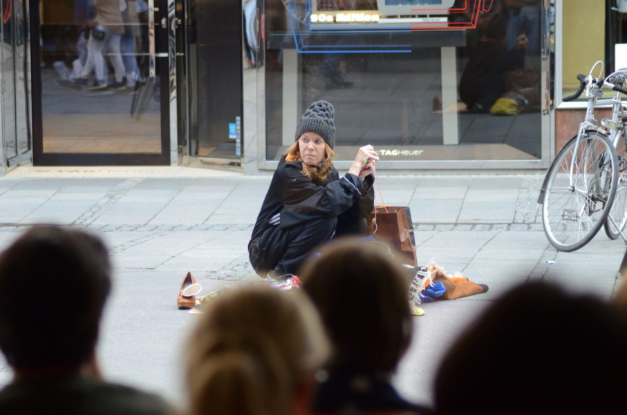 A woman in black clothing and a cap kneels on the ground in a pedestrian zone, surrounded by a few belongings. The heads of a number of people can be seen from behind in the foreground.