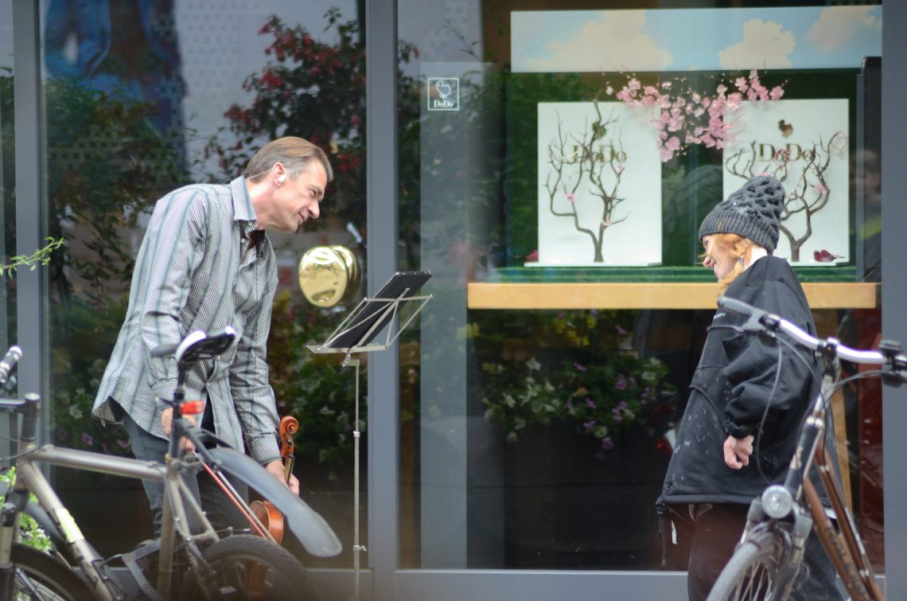 A woman dressed in black and wearing a black knitted hat and a man holding a violin in front of a music stand stand opposite each other in a shopping street, looking at each other. In the background is the display window of a jewellery shop.