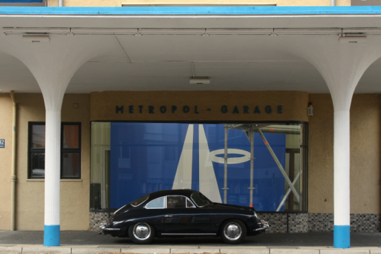 A black vintage Porsche is parked under the white and blue canopy of the former Metropol gas station.