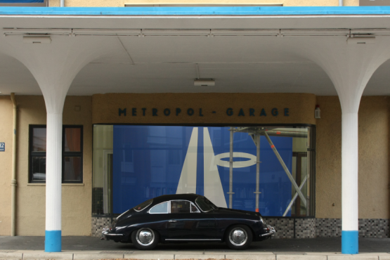 Ein schwarzer Porsche-Oldtimer ist unter dem weiß-blauen Vordach der ehemaligen Metropol Tankstelle geparkt.