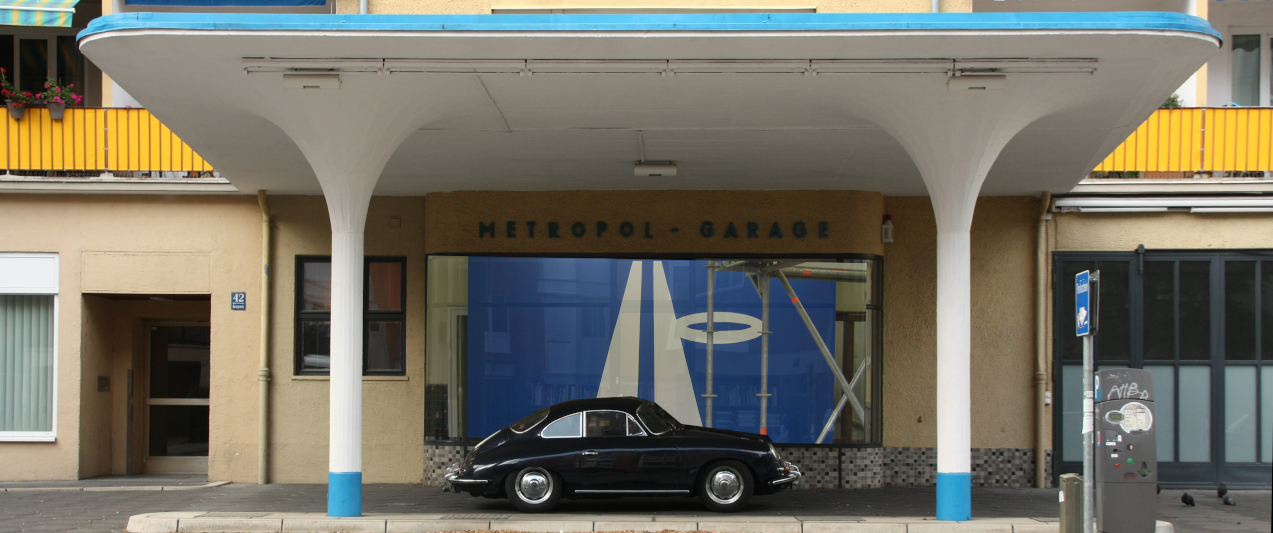 A black vintage Porsche is parked under the white and blue canopy of the former Metropol gas station.