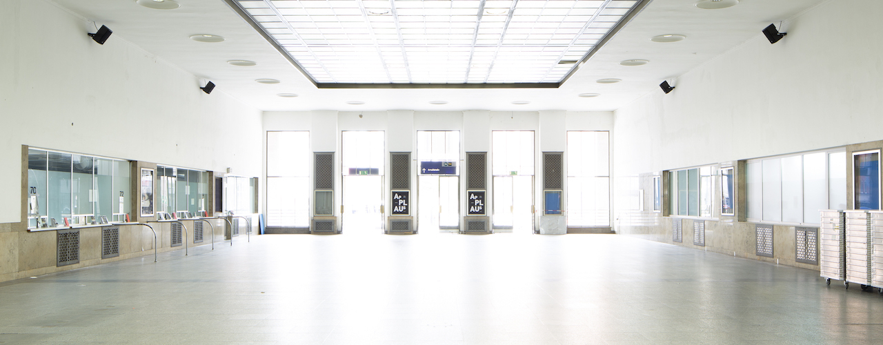 View of the empty station hall of the Starnberger Flügelbahnhof Munich with closed ticket counters. At the front of the concourse, between the exit doors, are black announcement posters with the project title "Applaus" in white lettering on a black background.