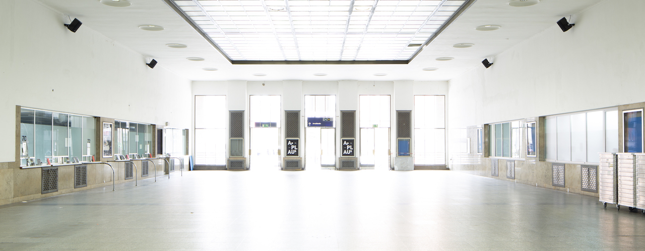 Blick in die leere Bahnhofsvorhalle des Starnberger Flügelbahnhofs mit geschlossenen Ticketschaltern. Am Kopfende der Halle, zwischen den Ausgangstüren, hängen schwarze Ankündigungsplakate mit dem Projekttitel "Applaus" in weißer Schrift auf schwarzem Grund.