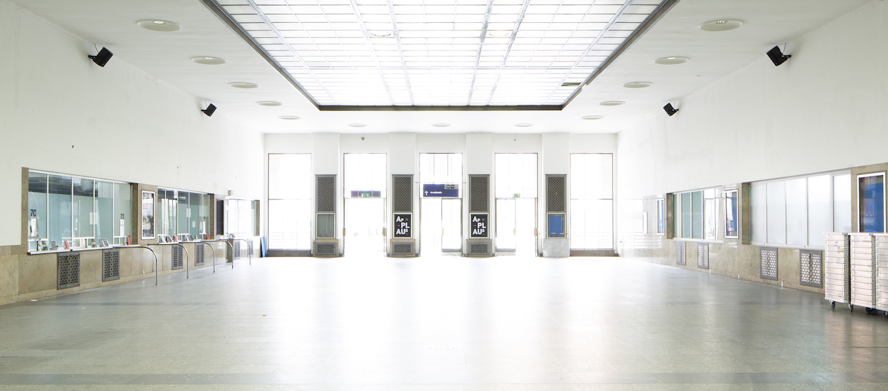 View of the empty station hall of the Starnberger Flügelbahnhof Munich with closed ticket counters. At the front of the concourse, between the exit doors, are black announcement posters with the project title "Applaus" in white lettering on a black background.