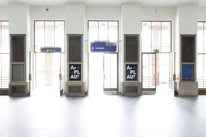 Blick auf den Ausgang der Bahnhofsvorhalle des Starnberger Flügelbahnhofs. Zwischen den Ausgangstüren hängen schwarze Ankündigungsplakate mit dem Projekttitel "Applaus" in weißer Schrift auf schwarzem Grund.