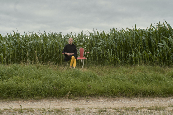 Im Hintergrund zu sehen ist ein Maisfeld vor wolkigem Himmel. Davor steht eine weiße Metallbank mit roten Polstern, darauf sitzend eine blonde Frau mit schwarzem Kleid und gelber Strumpfhose.
