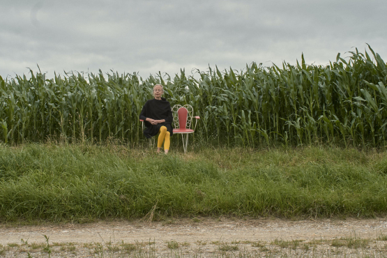 In the background is a cornfield against a cloudy sky. In front of it is a white metal bench with red cushions, on which sits a blonde woman in a black dress and yellow tights.