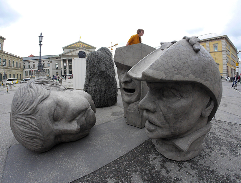 Zu sehen sind drei graue Skulpturen in Form überdimensionierter Köpfe, die auf dem Max-Joseph-Platz stehen. Die Köpfe zeigen die Konterfeis von Angela Merkel, EZB-Chef Mario Draghi und Dutty Boukman, dem Anführer des ersten Sklavenaufstandes 1791 in Haiti.