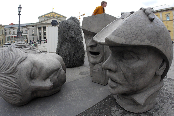 Zu sehen sind drei graue Skulpturen in Form überdimensionierter Köpfe, die auf dem Max-Joseph-Platz stehen. Die Köpfe zeigen die Konterfeis von Angela Merkel, EZB-Chef Mario Draghi und Dutty Boukman, dem Anführer des ersten Sklavenaufstandes 1791 in Haiti.