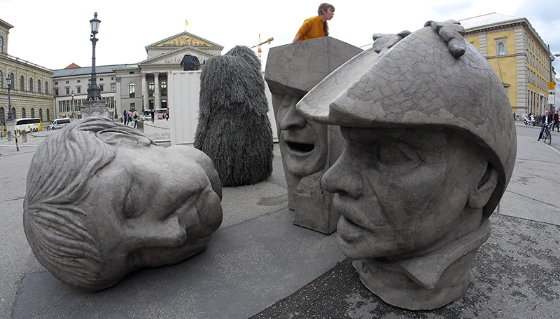 Zu sehen sind drei graue Skulpturen in Form überdimensionierter Köpfe, die auf dem Max-Joseph-Platz stehen. Die Köpfe zeigen die Konterfeis von Angela Merkel, EZB-Chef Mario Draghi und Dutty Boukman, dem Anführer des ersten Sklavenaufstandes 1791 in Haiti.