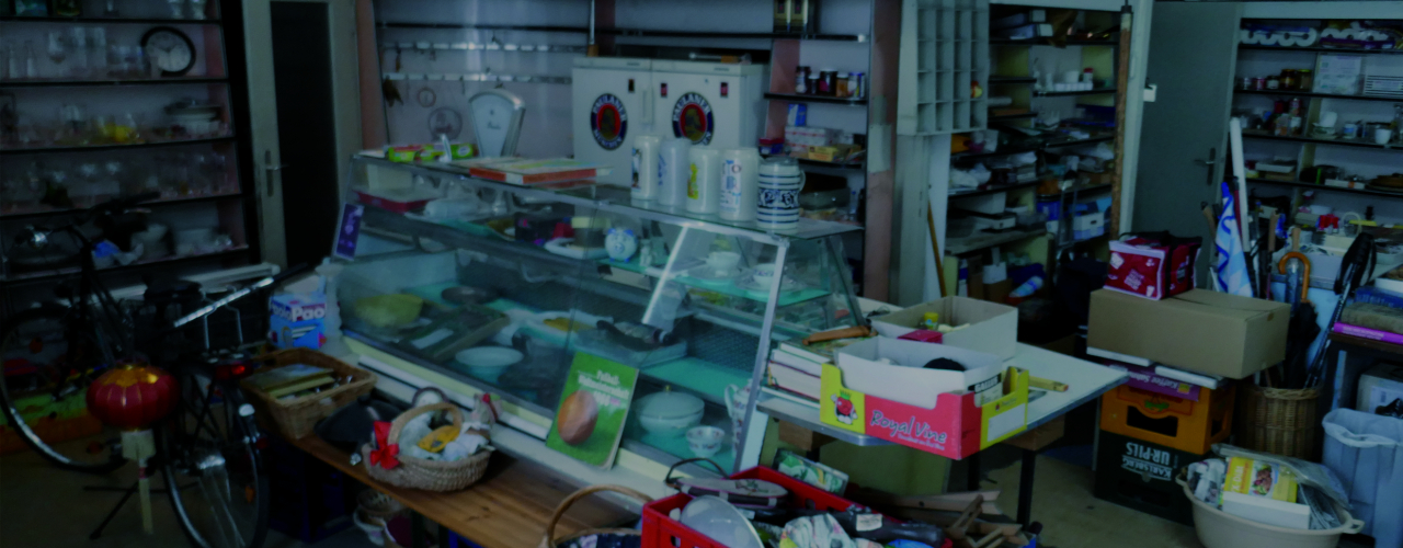 View of an abandoned grocer's shop with a counter in the middle. The shop is filled with discarded household items and the old range of goods on the shelves.