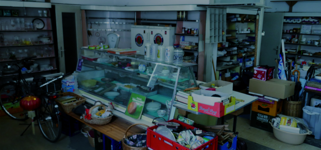 View of an abandoned grocer's shop with a counter in the middle. The shop is filled with discarded household items and the old range of goods on the shelves.