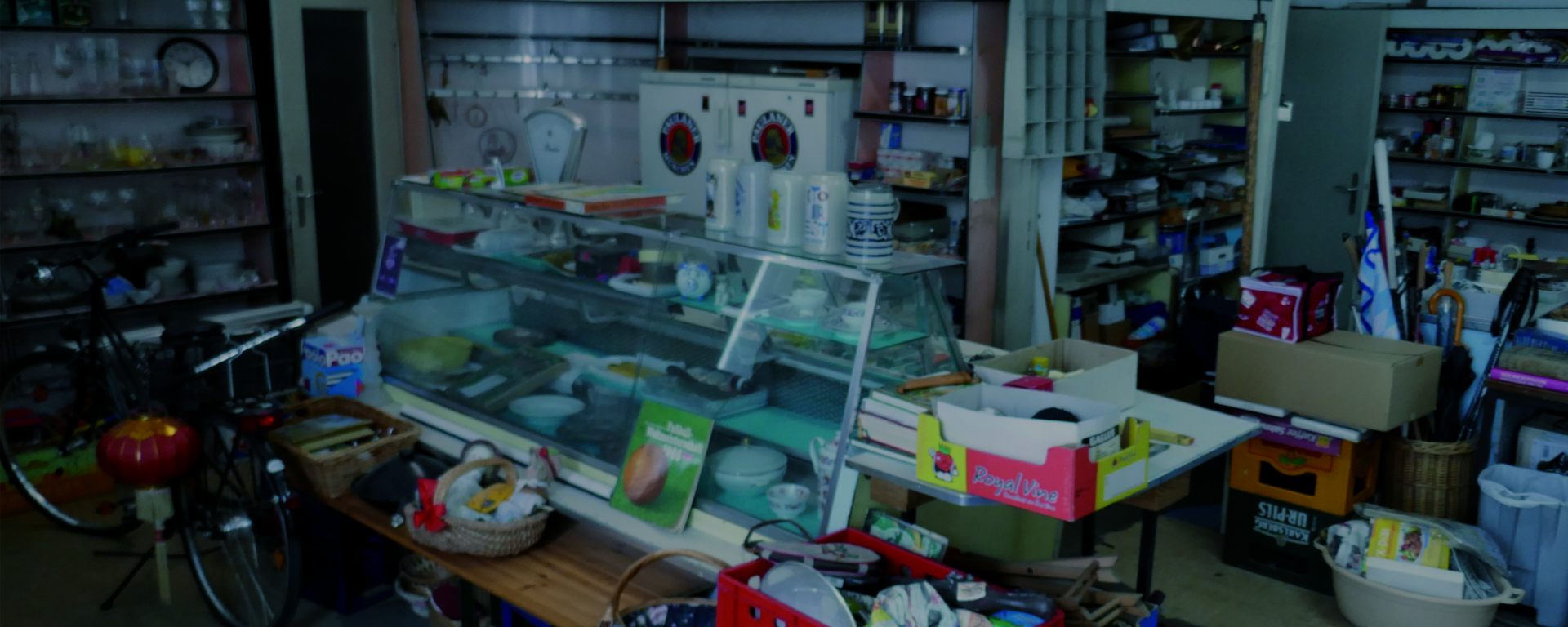View of an abandoned grocer's shop with a counter in the middle. The shop is filled with discarded household items and the old range of goods on the shelves.
