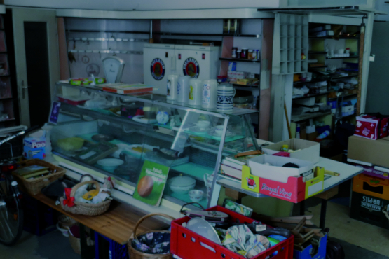 View of an abandoned grocer's shop with a counter in the middle. The shop is filled with discarded household items and the old range of goods on the shelves.