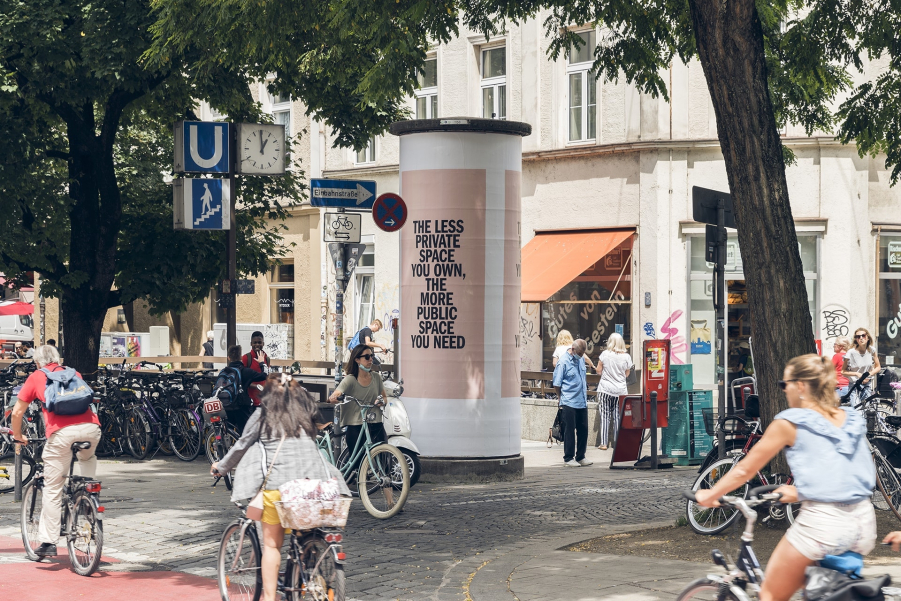 Street scene with passers-by. In the centre of the picture is an advertising pillar next to the entrance of an underground station. The advertising pillar reads "THE LESS PRIVATE SPACE YOU OWN, THE MORE PUBLIC SPACE YOU NEED" on a pink background.