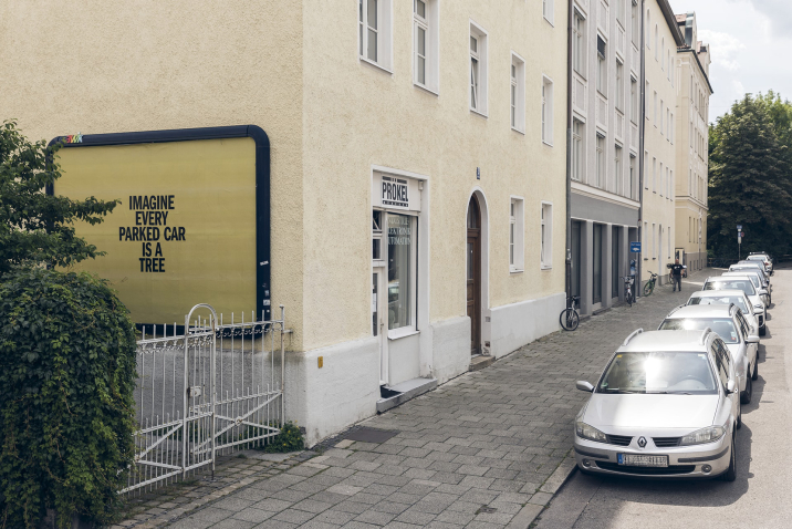 Ansicht einer Häuserreihe mit vor dem Gehsteig geparkten Autos. Auf der Hauswand einer Einfahrt hängt eine Werbefläche, darauf zu lesen ist auf gelbem Hintergrund der Satz: "IMAGINE EVERY PARKED CAR IS A TREE".