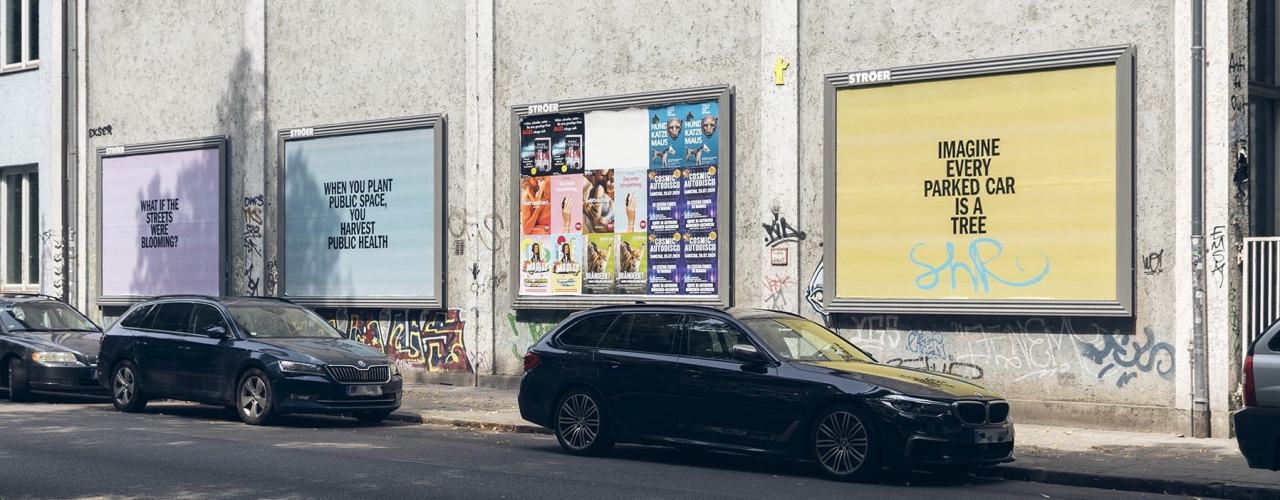 View of several colourful advertising billboards mounted on a house wall with black text on them. In front, you see a row of parked cars.