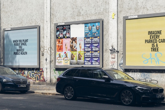 View of several colourful advertising billboards mounted on a house wall with black text on them. In front, you see a row of parked cars.
