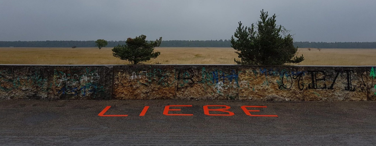 In front of a wall, the word "Liebe" ("Love") is taped to the ground in red capital letters. In the background is a brown field and beyond that the edge of a forest.