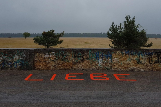 In front of a wall, the word "Liebe" ("Love") is taped to the ground in red capital letters. In the background is a brown field and beyond that the edge of a forest.