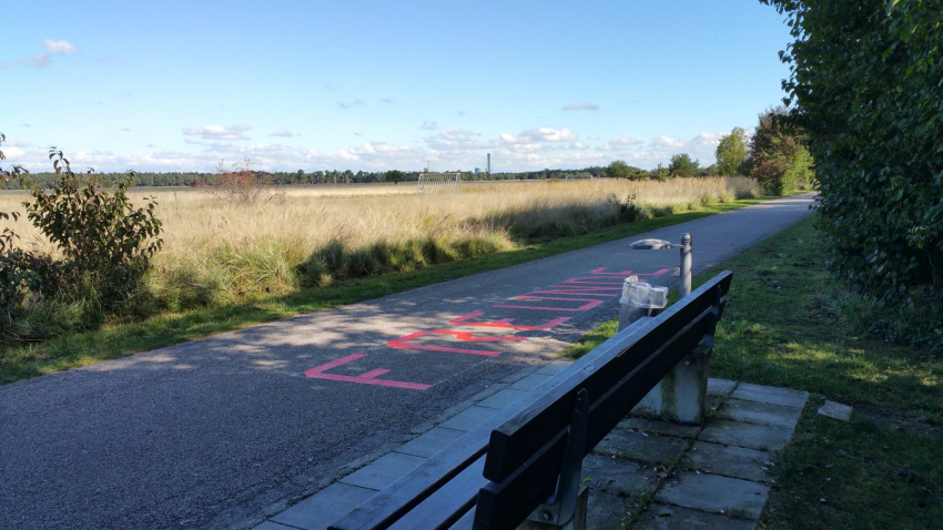 Ansicht eines Gehweges am Rande eines Feldes. Neben dem Weg eine Bank mit Mülleimer. Auf den Weg ist in roten Großbuchstaben das Wort "Freunde" aufgeklebt.