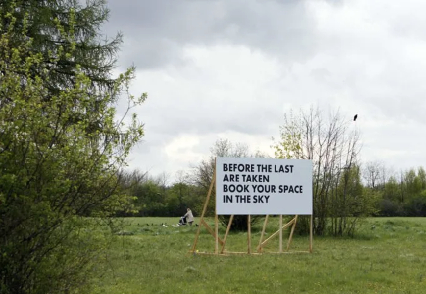 Weiße Reklametafel mit einem Holzgestell auf einer Wiese mit Bäumen. In schwarzer Schrift in Großbuchstaben zu lesen: "Before the last are taken, book your space in the sky".