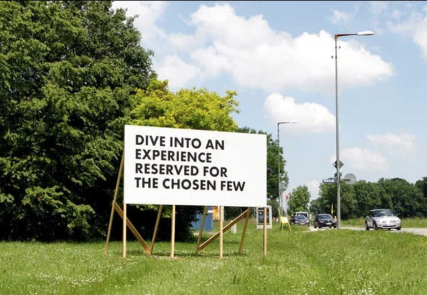 Weiße Reklametafel mit einem Holzgestell auf einer Wiese neben einer Straße mit vorbeifahrenden Autos. In schwarzer Schrift in Großbuchstaben zu lesen: "Dive into an experience reserved for the chosen few".