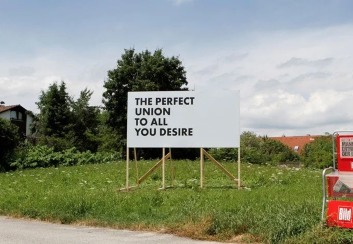 Weiße Reklametafel mit einem Holzgestell auf einer Wiese in einem Wohngebiet. In schwarzer Schrift in Großbuchstaben zu lesen: "The perfect union to all you desire".