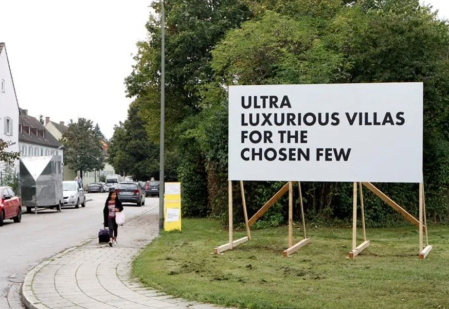 Weiße Reklametafel mit einem Holzgestell auf einer Wiese in einem Wohngebiet. In schwarzer Schrift in Großbuchstaben zu lesen: "Ultra luxurious villas for the chosen few".