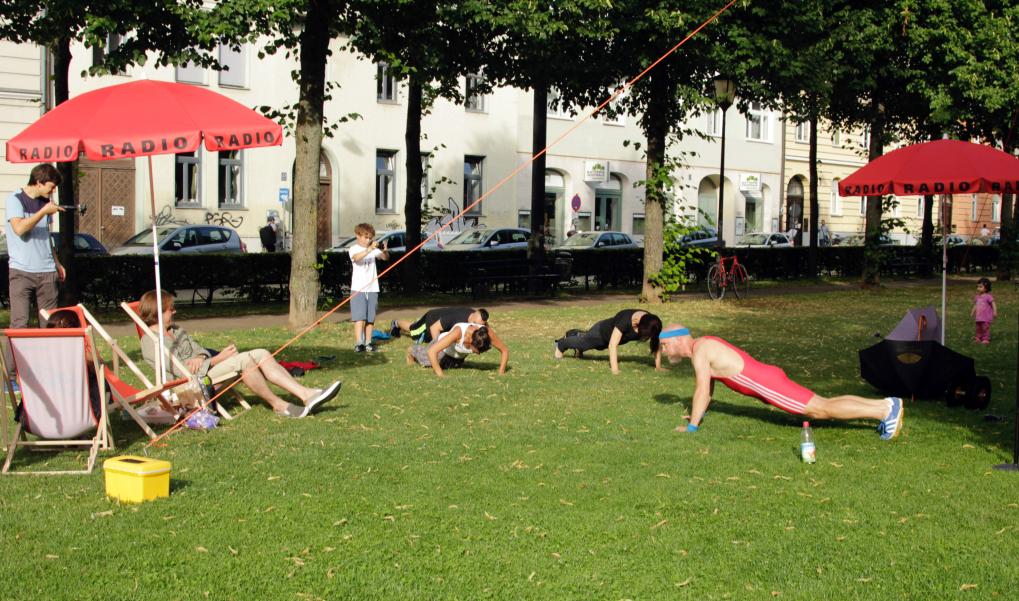 View of Bordeauxplatz with magenta-colored parasols with the word "Radio" on them and deckchairs. In between, several people in sports outfits are doing press-ups.